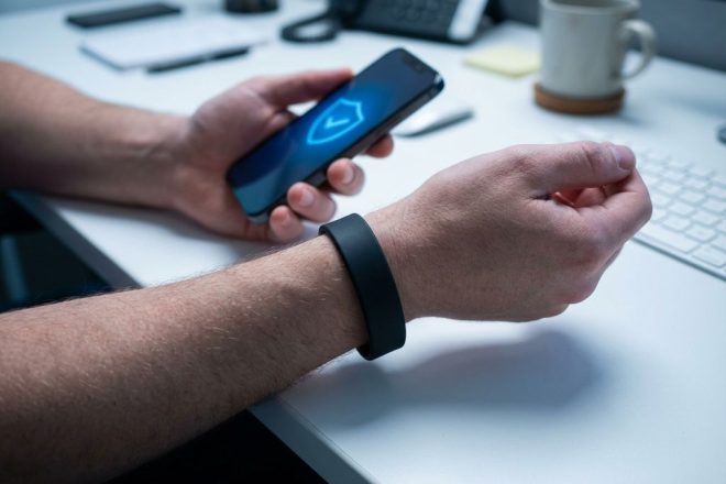 Persoon draagt zwarte RFID-polsband terwijl smartphone met beveiligingspictogram erboven wordt gehouden op wit bureau